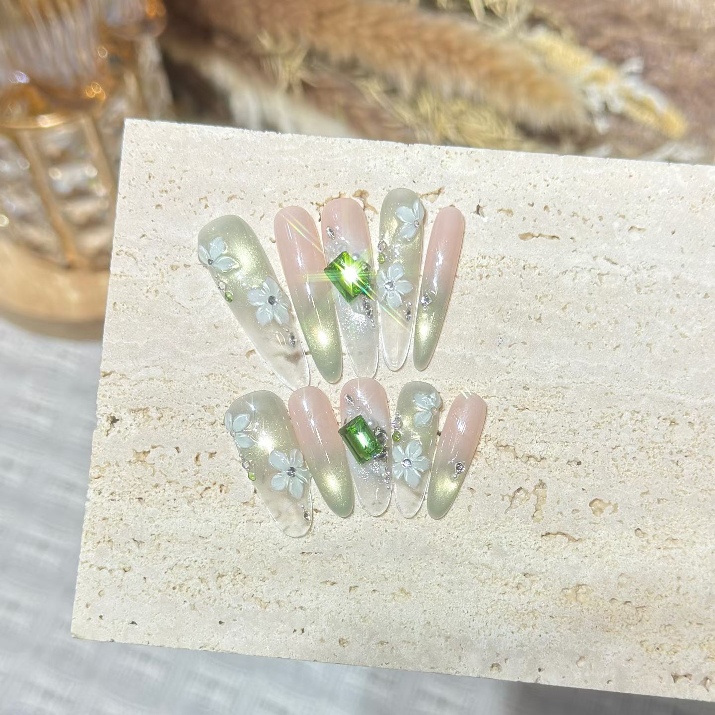 Pale green and blush press-on nail set with floral decoration and green gems, shown on a stone display